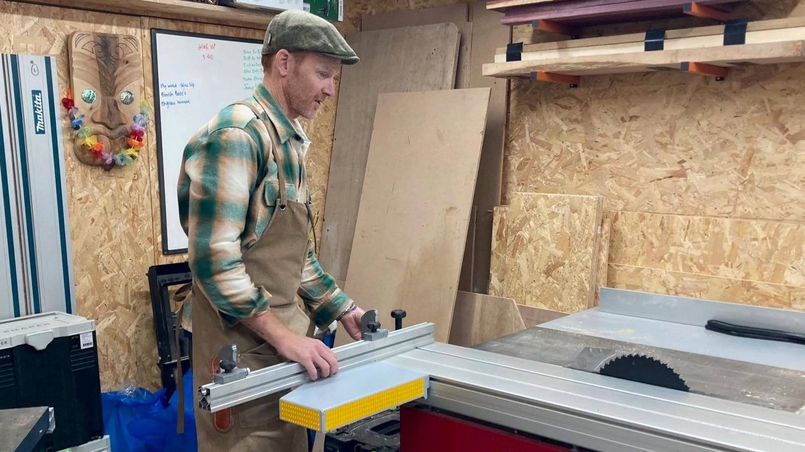 Steve Bate using measuring tools in a carpentry workshop wearing a flat cap, green checked shirt and brown carpentry apron
