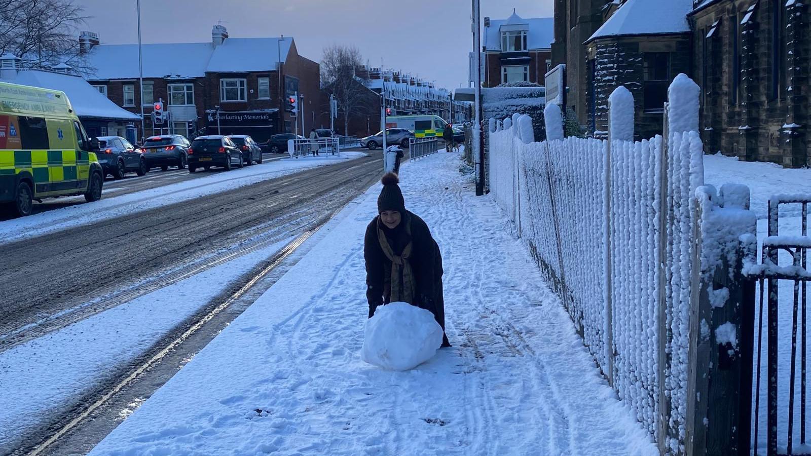 A woman is pushing a snowball, which comes up to her knees, along a pavement. The road is slushy with ice behind her.