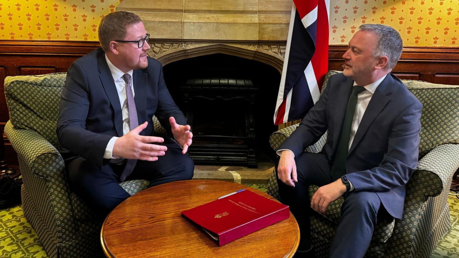Jonathan Brash sits in a green armchair, wearing a navy suit and purple tie and glasses, holding his hands out over a table. Opposite him in a similar armchair is Steve Reed, looking at Jonathan Brash, wearing a navy suit and white tie.