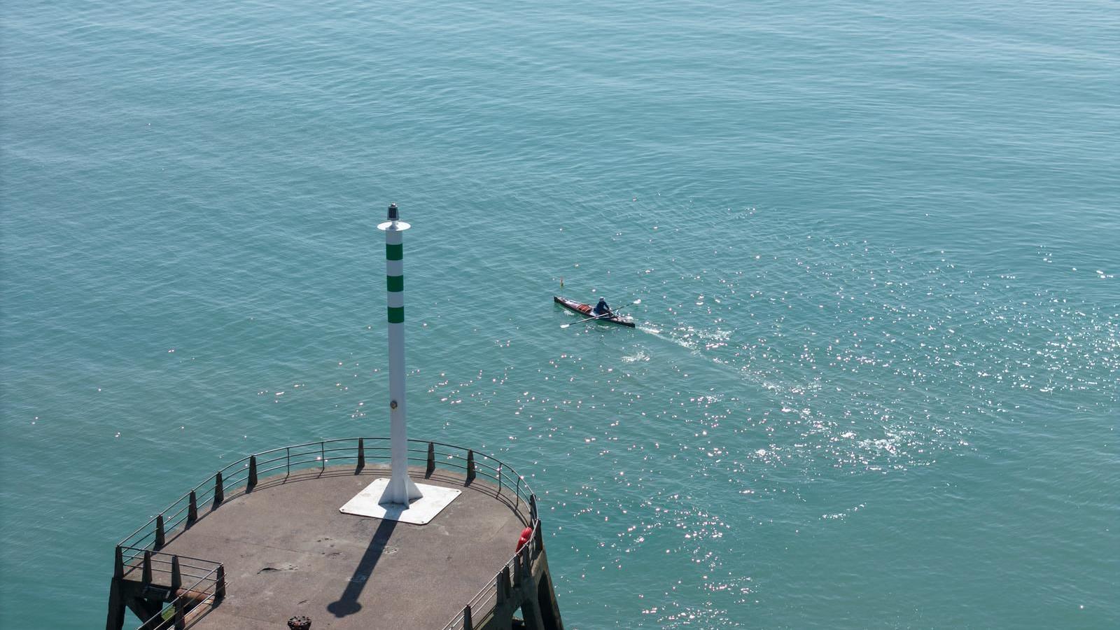 An aerial view showing a rowing boat in the sea with the sun reflecting off the still water. In the foreground is a white and green pole on top of a metal platform.