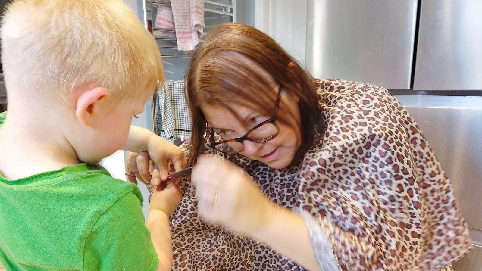 Sam, with short brown hair and wearing a leopard-print top, sitting on a chair and helping a young boy with blonde hair and wearing a green t-shirt cut her hair with scissors.