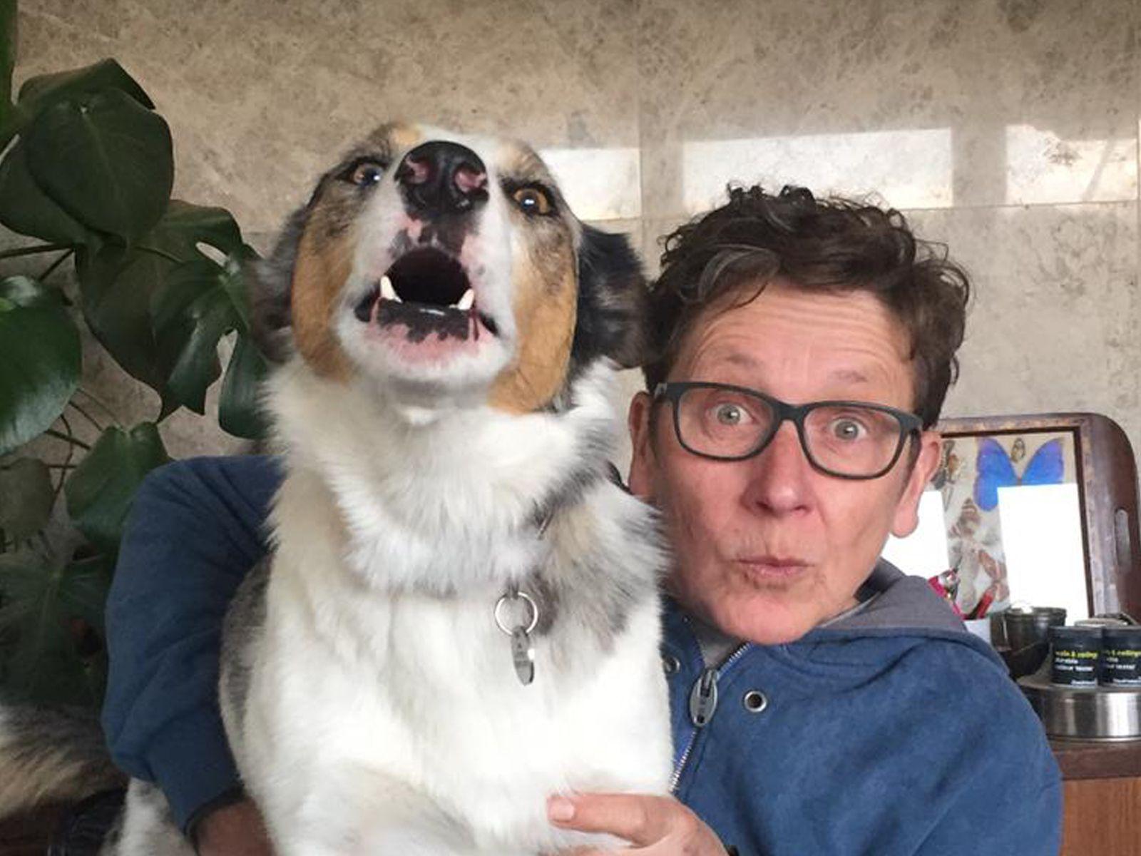 A woman and a dog stand together inside a living room. The woman has short brown hair and glasses, and is wearing a blue hoodie. The dog has white and brown fur. The room behind them contains a plant and a sideboard.