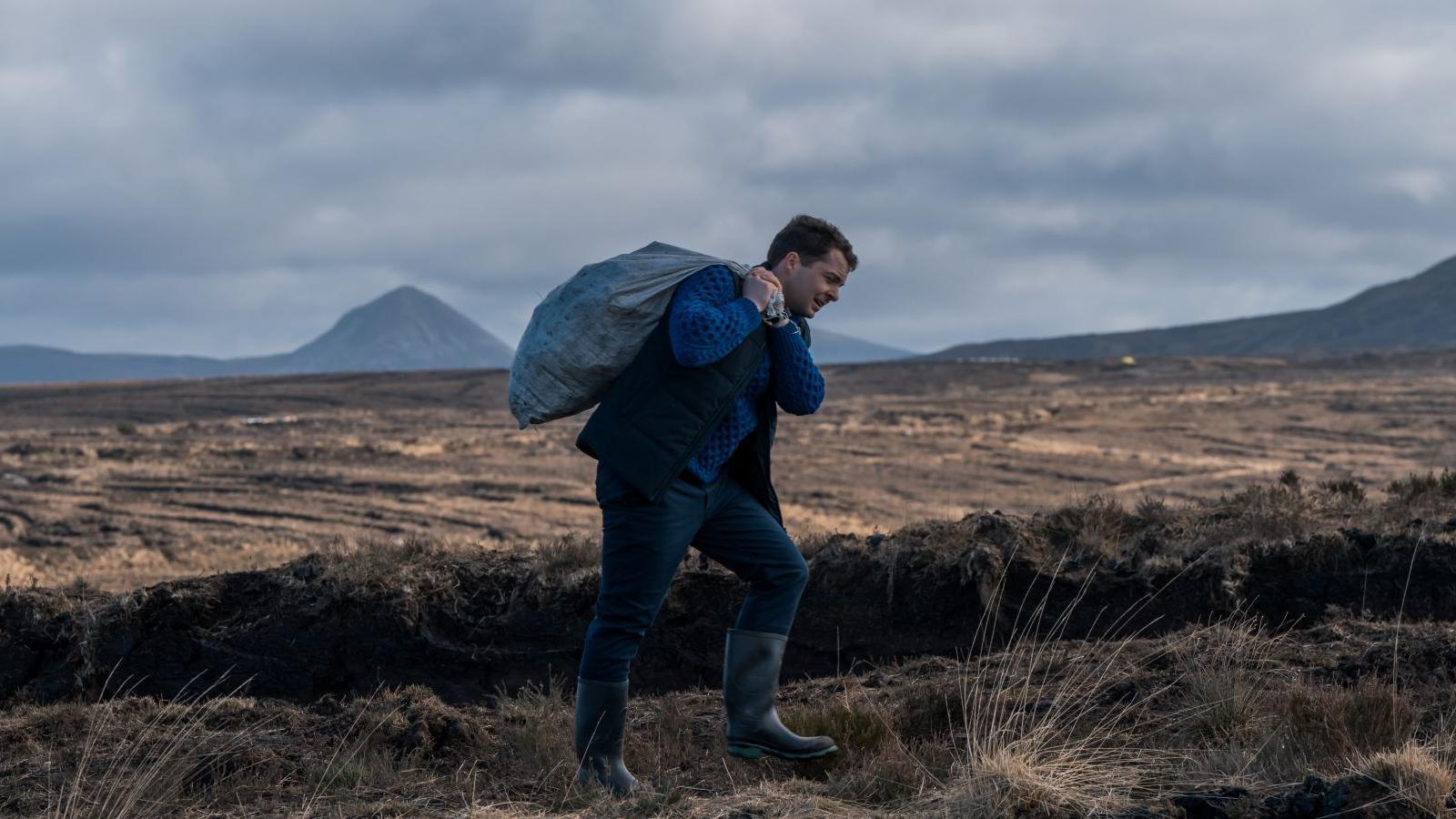 Alex Murphy wearing a blue fisherman's jumper, a dark gilet, trousers and wellie boots is carrying a heavy sack over his shoulder as he walks through an empty bog land.