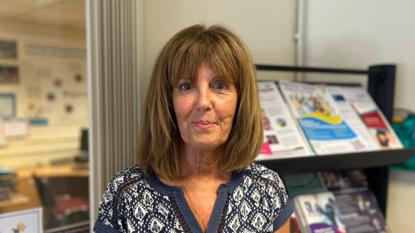 A woman with shoulder length brown hair is looking into the camera. She is wearing a blue and white patterned top. The background is out if focus but she is standing in front of an information stand with a number of leaflets and booklets.