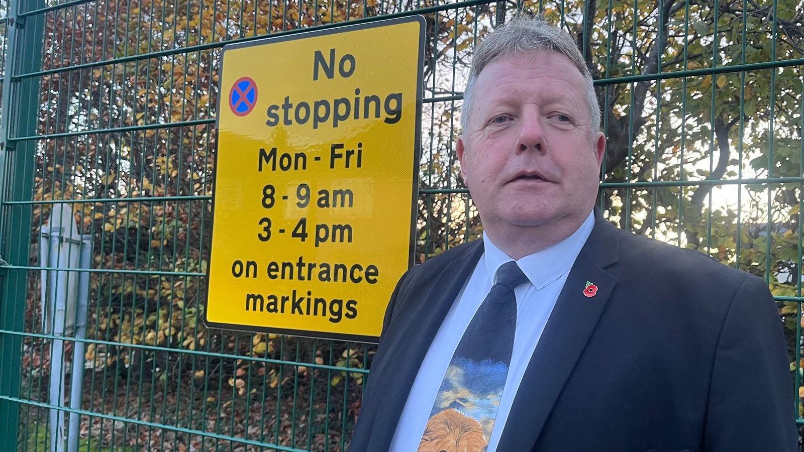 A smartly dressed man with grey hair stands in front of a tall, green wire fence. He is wearing a navy blue suit, light blue shirt, a blue tie with a painted lion on it, and a poppy lapel badge. Attached to the fence is a large yellow sign. It reads: "Not stopping, Mon-Fri, 8-9am, 3-4pm, on entrance markings." Trees with autumn leaves stand in the background.