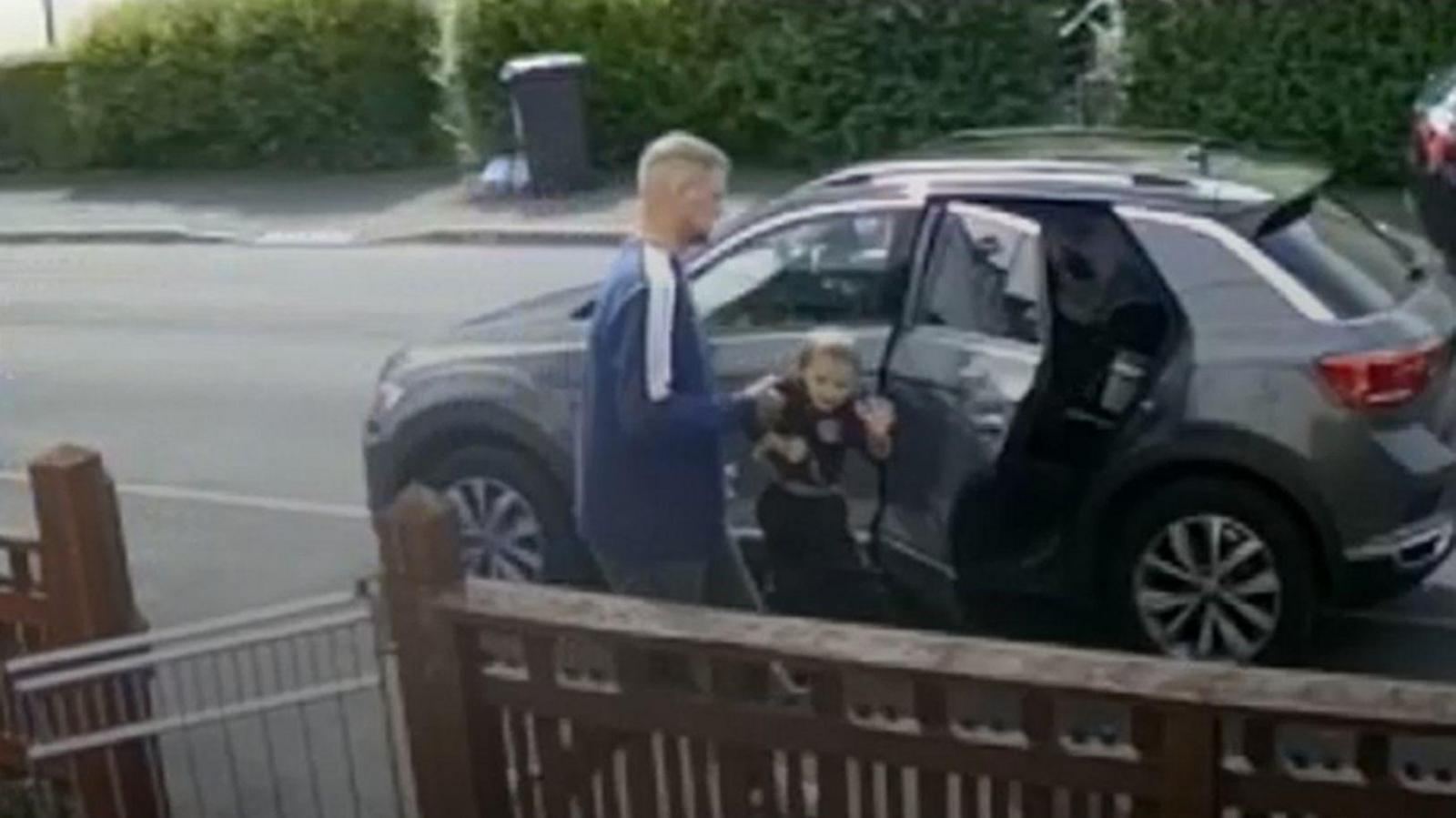 A man grabs a child by his arm as they exit a black car. In the foreground you can see a gate attached to a wooden fence leading to a house.