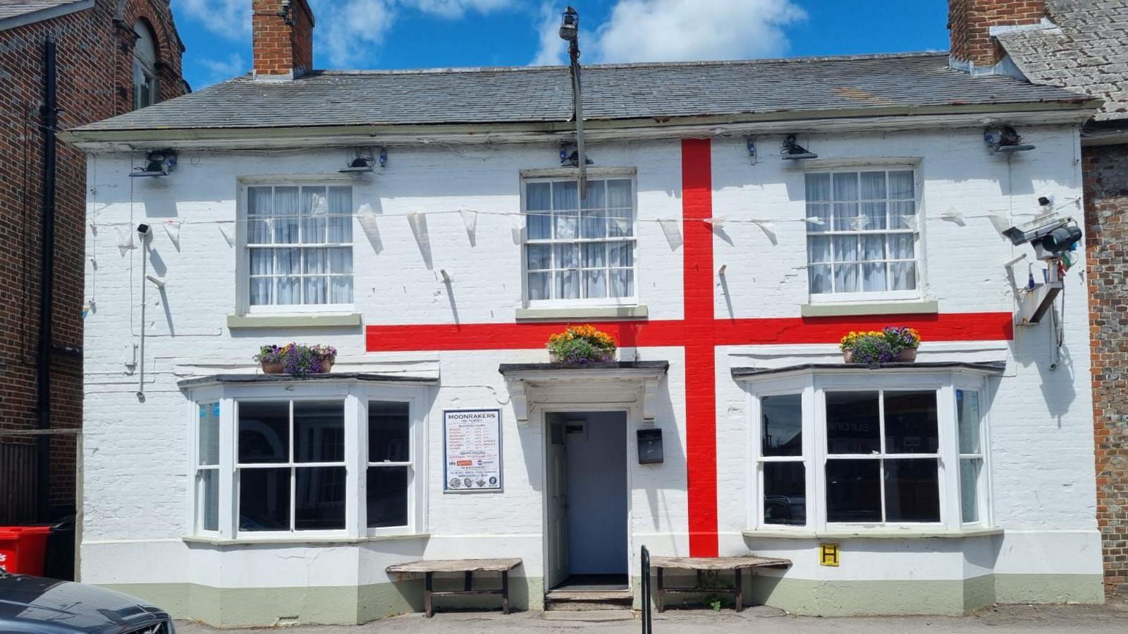 A white pub building with five windows, a CCTV camera and a large red cross painted on the exterior