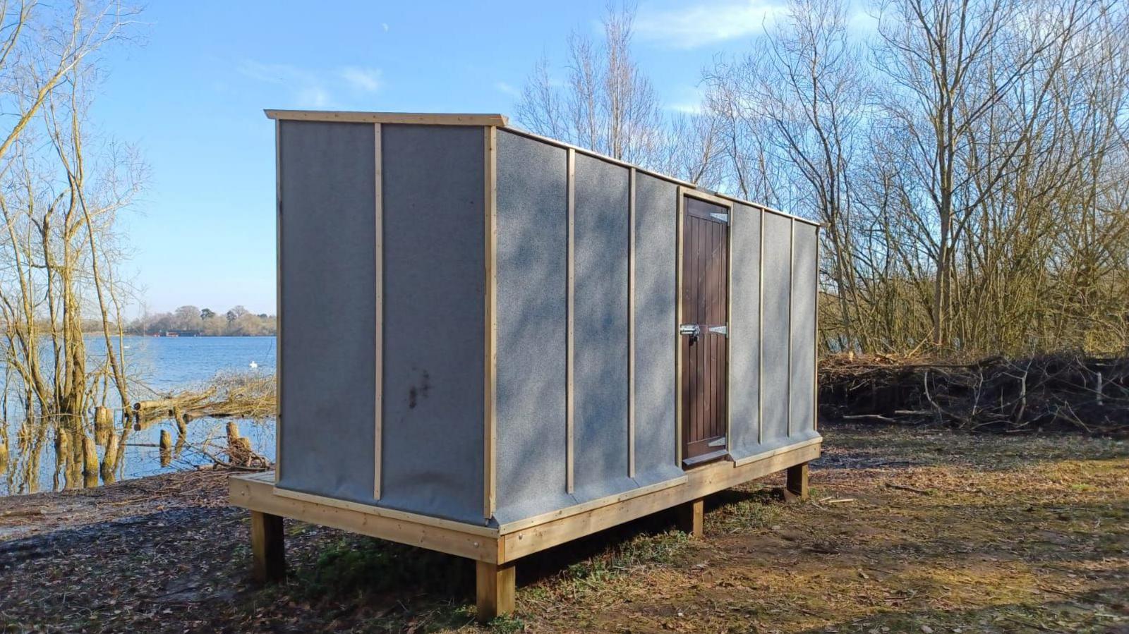 Tall wooden box divided into sections, standing on a wooden platform on a dirt surface with the blue-coloured reservoir to the left. Trees are visible in the distance.