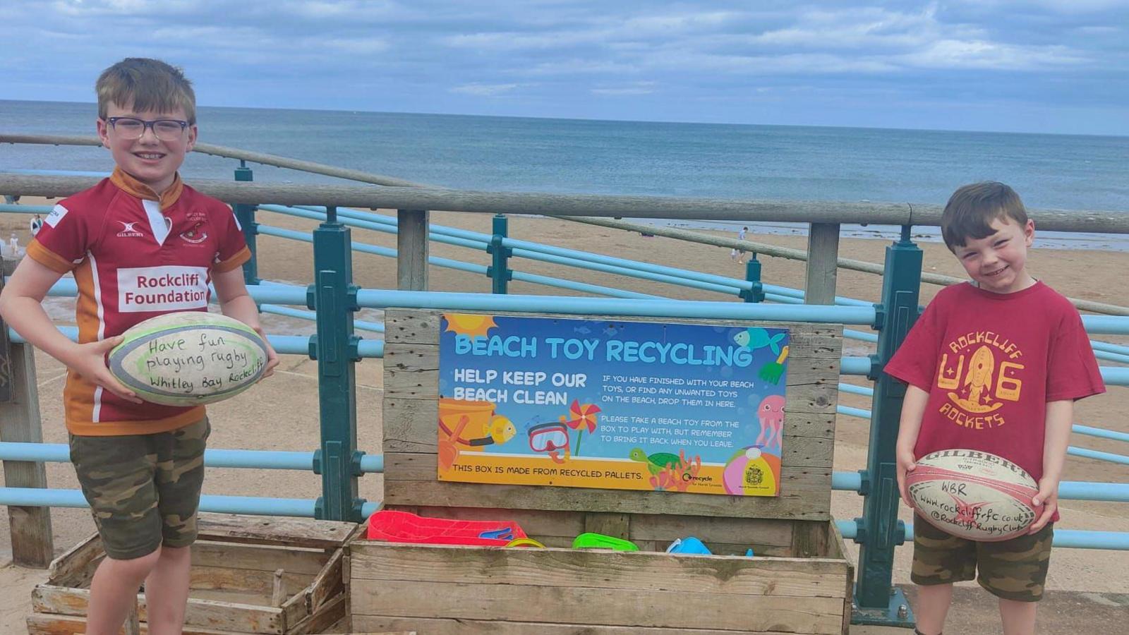 Benjamin and Alexander both holding rugby balls at Longsands beach. They are wearing red Rockcliffe Rugby club shorts and green camouflage shorts. They are standing between a wooden chest, where people can donate toys for other people to use.