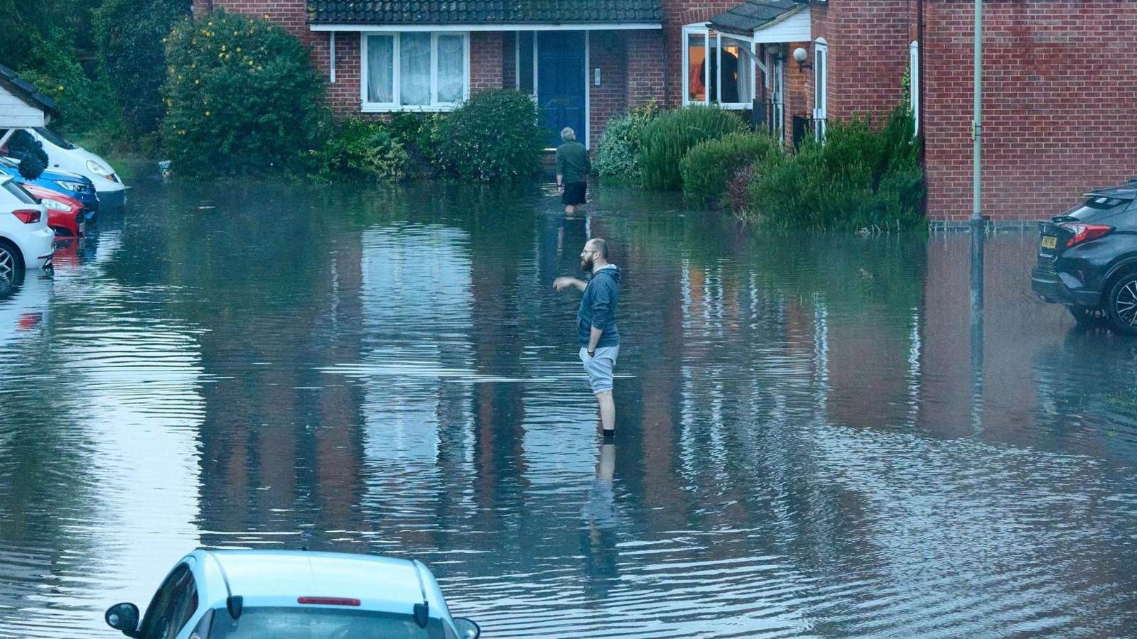 View from a window on to a flooded road in Cheltenham. Two men are standing ankle-deep in the water, assessing the damage.