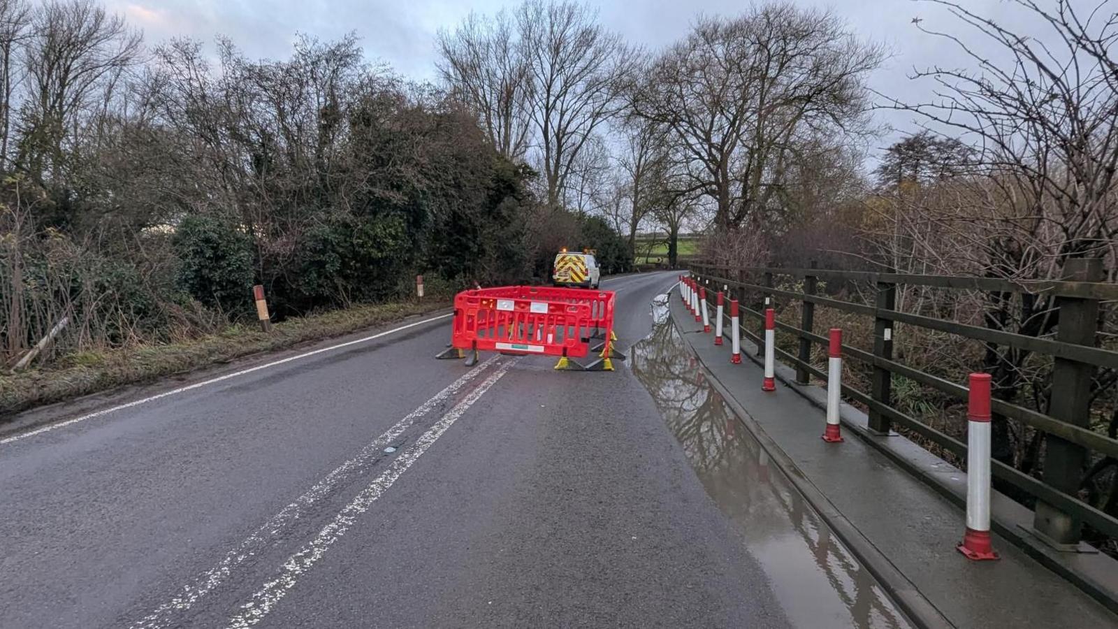 The A361 with a large cordoned of area in the middle. The road is above a water course, and their is a yellow and red van parked in the near distance.