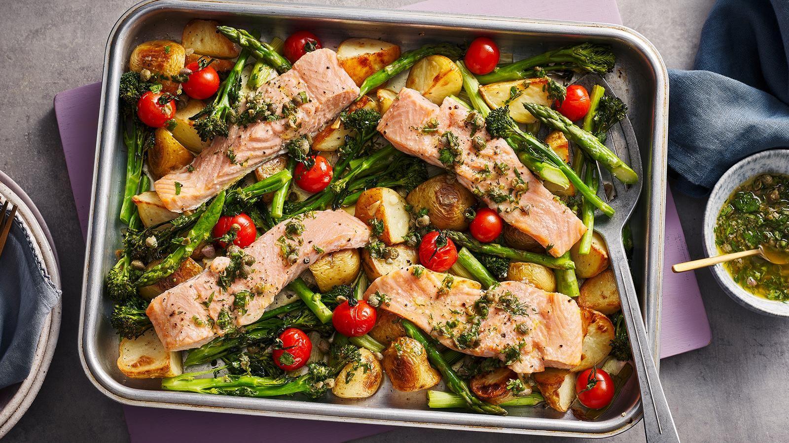A silver roasting tray filled with chopped asparagus, potatoes, tomatoes and fillets of salmon, placed on a purple board. 