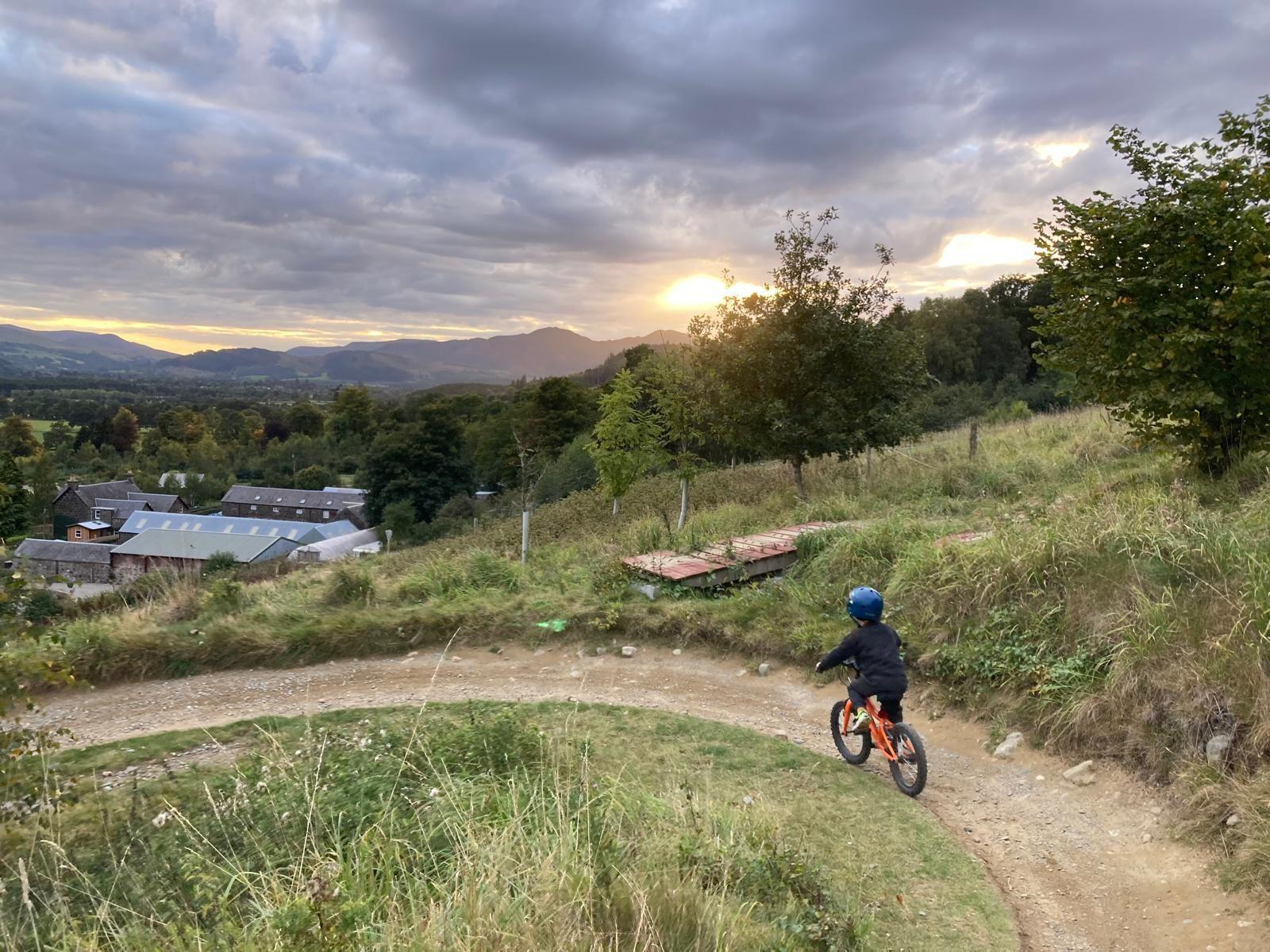 A young boy, on his bike and wearting a cycling helmet, takes a bend on a downhill cycle path with a wonderful view of a village, trees, rolling hills and the sun rising in the sky beyond