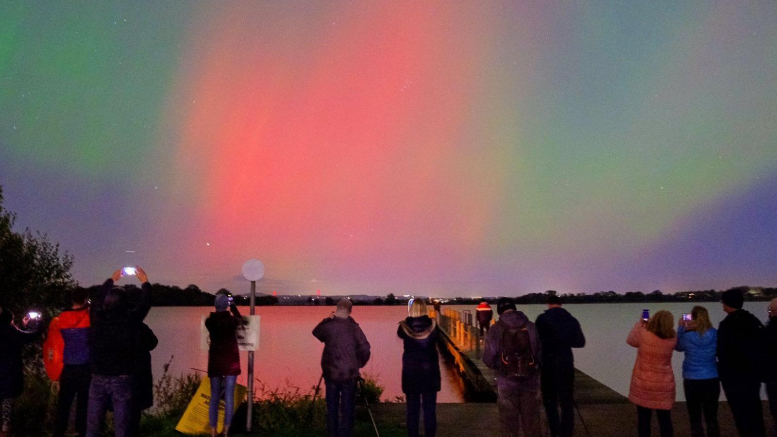 Green and red beams of light are visible in a purple sky with stars above a lake. Below a number of people take photos on phones and cameras.