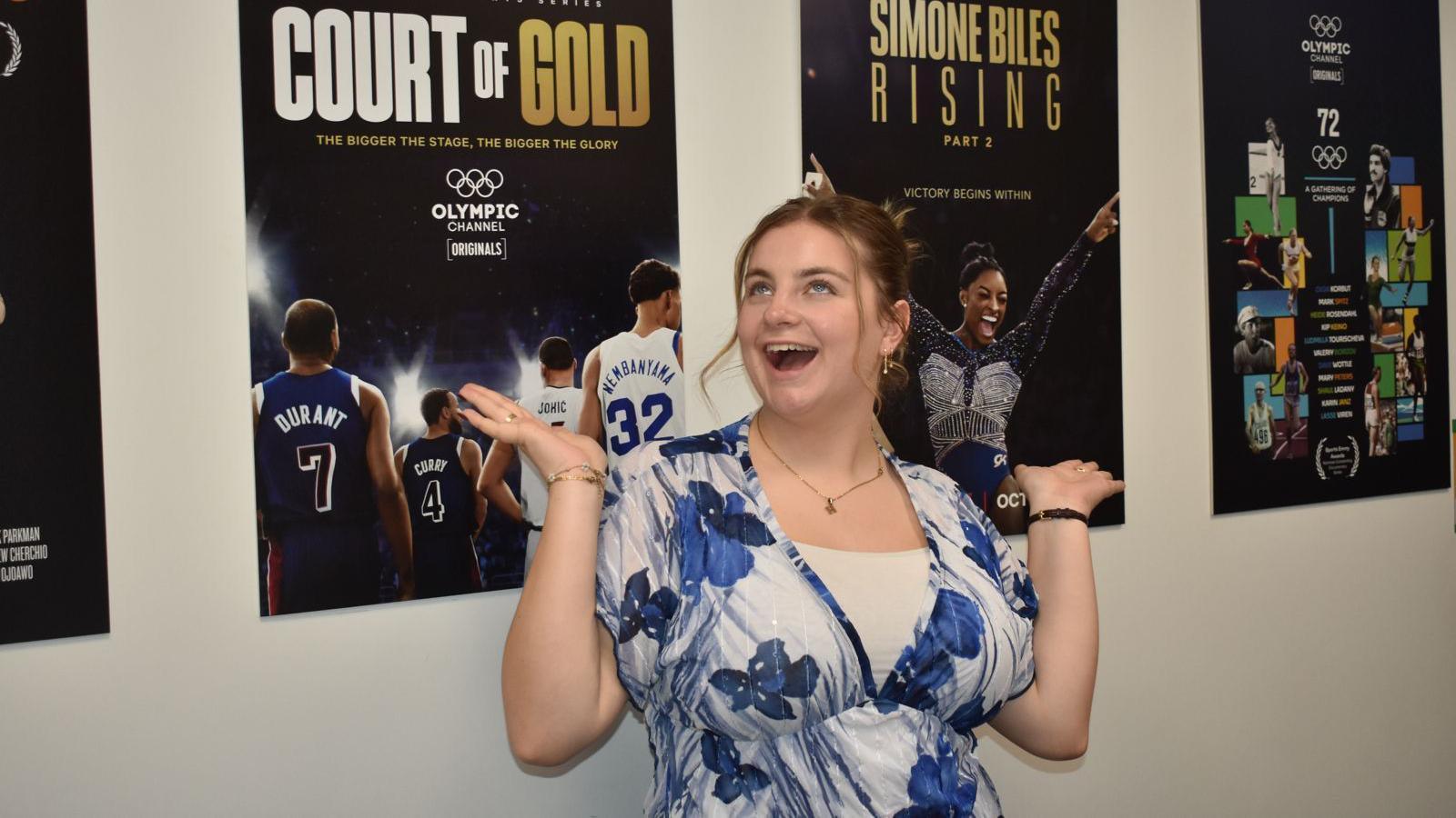 Holly Norman wearing a blue and white floral patterned blouse with her dark blonde hair tied back in a bun with small curled pieces pulled out at the front. She is smiling and looking upwards, posing for a picture with her hands in the air beside her. On the white wall behind her there are four large black posters of Olympic documentaries made by the channel, including one on Simone Biles and another on men's basketball.