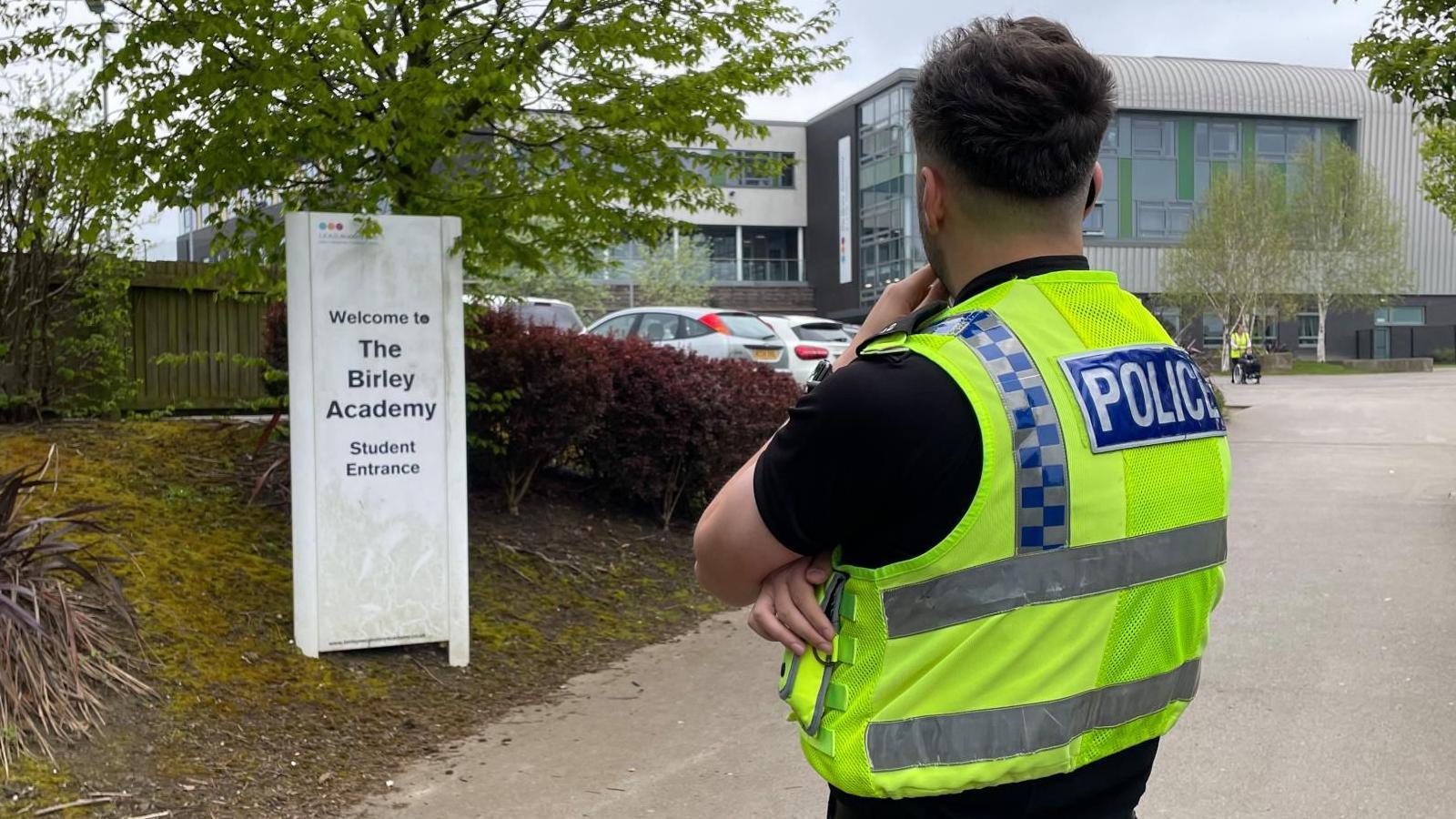 A police officer stands looking at a school building and car park, near a sign reading "The Birley Academy student entrance".