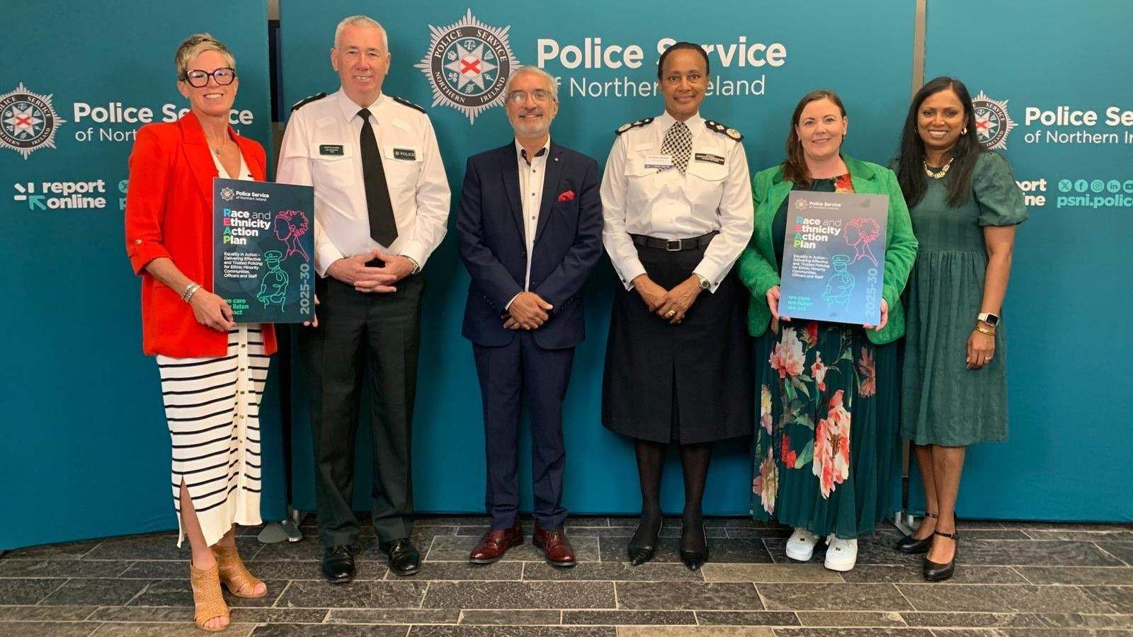 Jon Boutcher, along with five other people, standing in front of a large PSNI poster. All six are smiling at the camera, and the woman on the far left is holding a sign displaying the name of the strategy. 