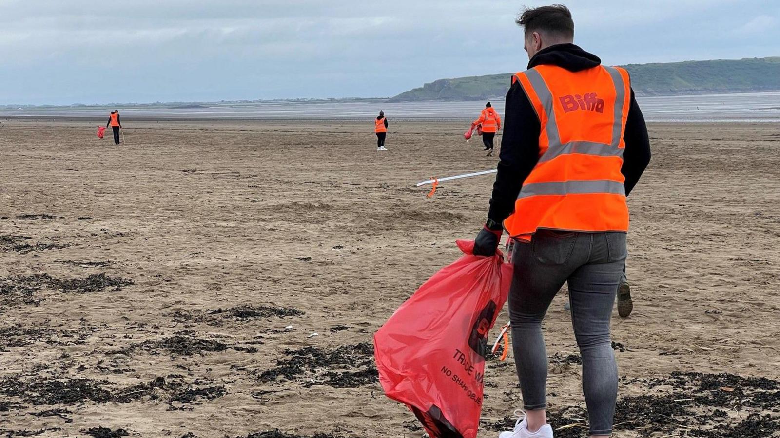 Volunteer in an orange hi viz Biffa jacket collects litter at a beach on an overcast day. He's facing away from the camera. Other volunteers can be seen ahead in the distance.