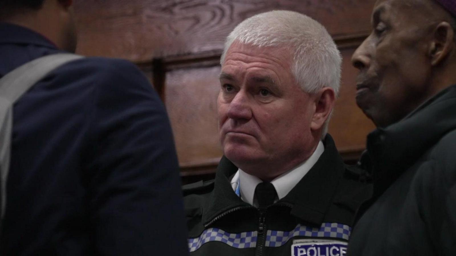 A man in a police uniform is talking to members of a church congregation. He has short white hair and is staring directly into the eyes of a man he is talking to.