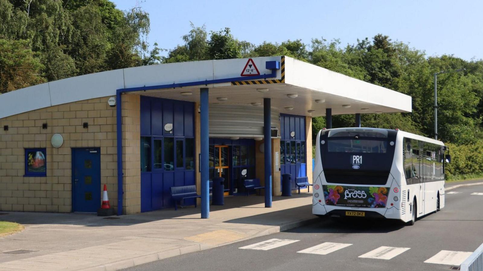 A white PR1 park and ride white bus is waiting at the bus shelter at Belmont. there is a seating area and bins.
