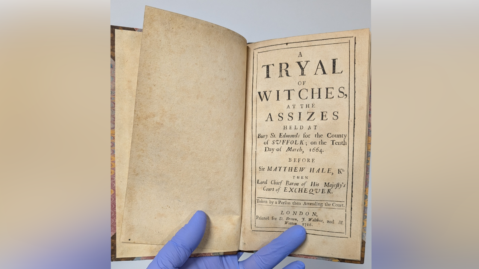 A historic book that recounts the Bury St Edmunds witch trials. It shows the first page of the book, which someone wearing blue gloves holds open.
