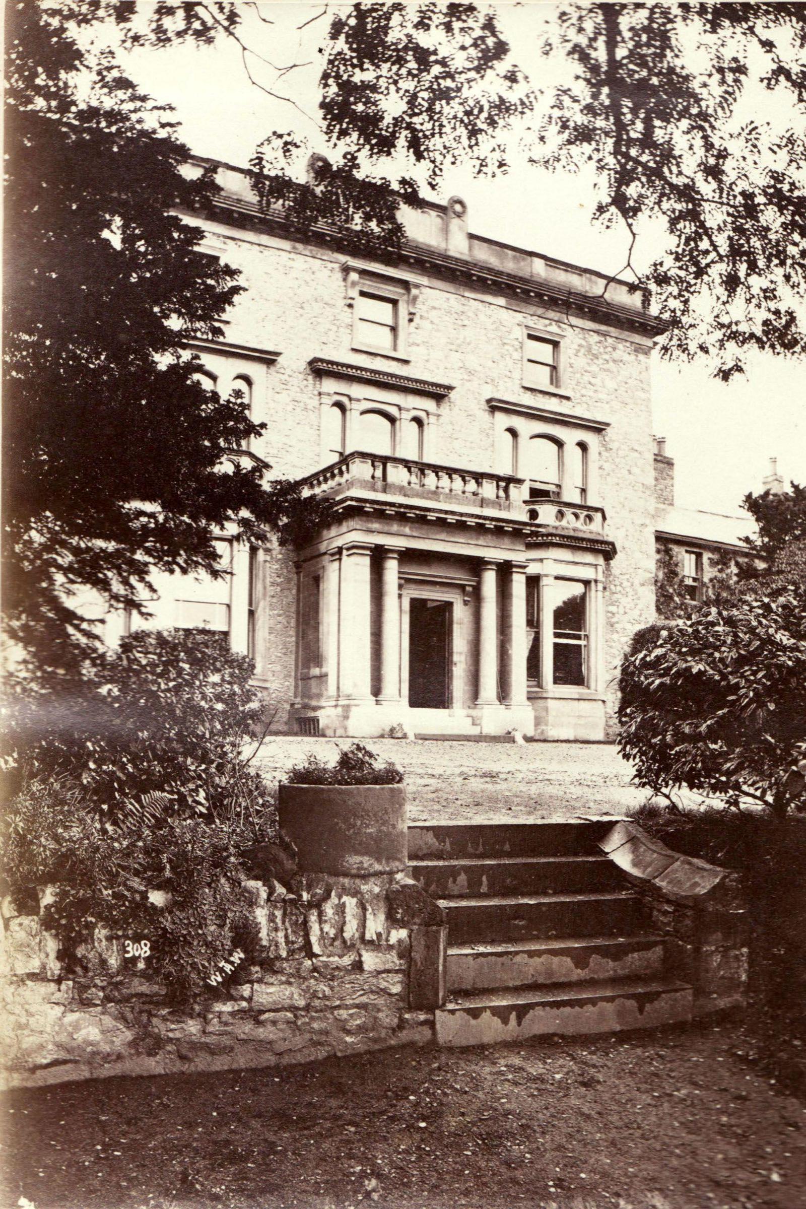The driveway leads to a porticoed entrance before a Georgian-style manor house. Steps lead down to a garden area.