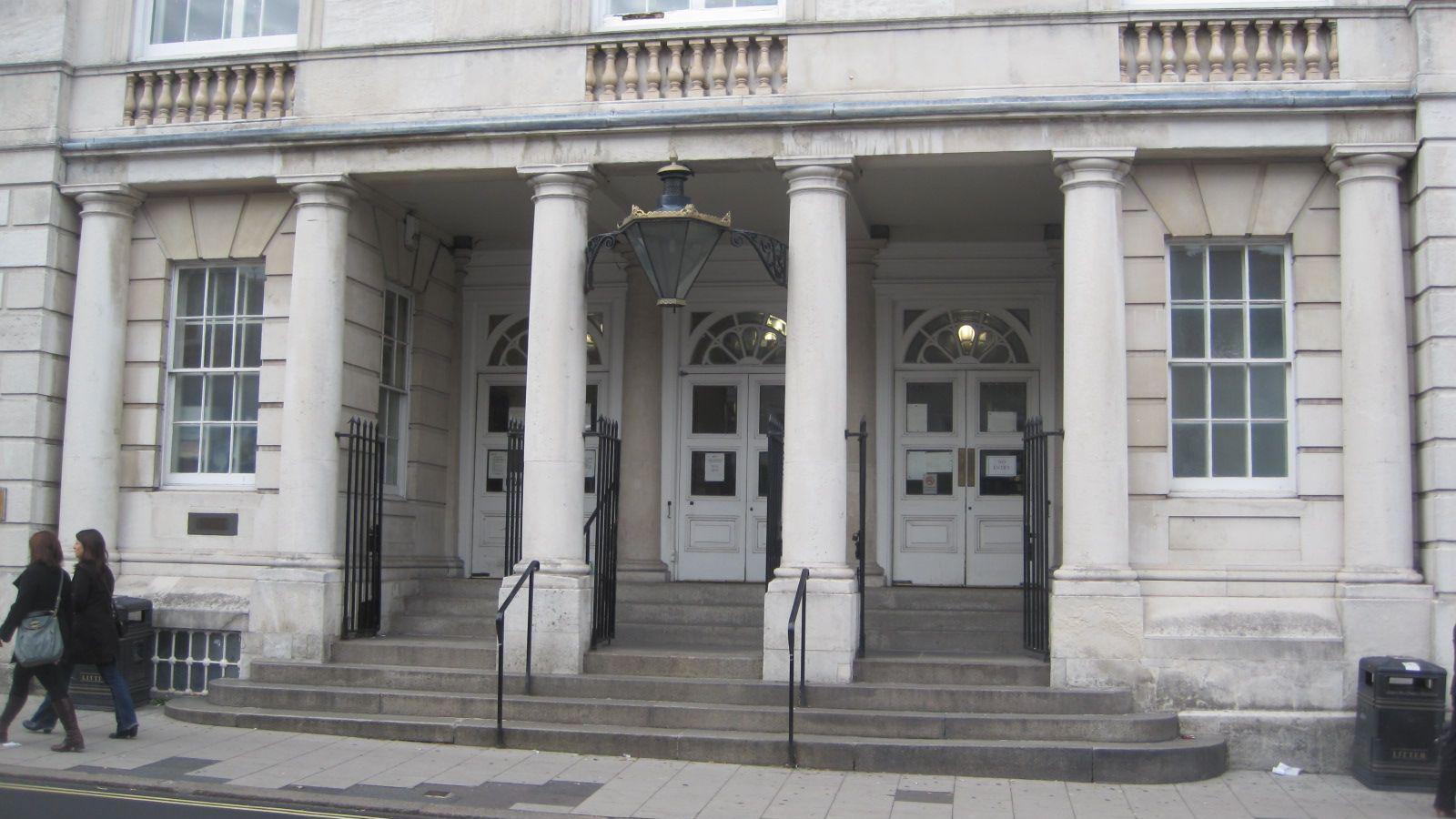 The exterior of a grey stone building. The image shows the steps and large pillar leading to three white doors.