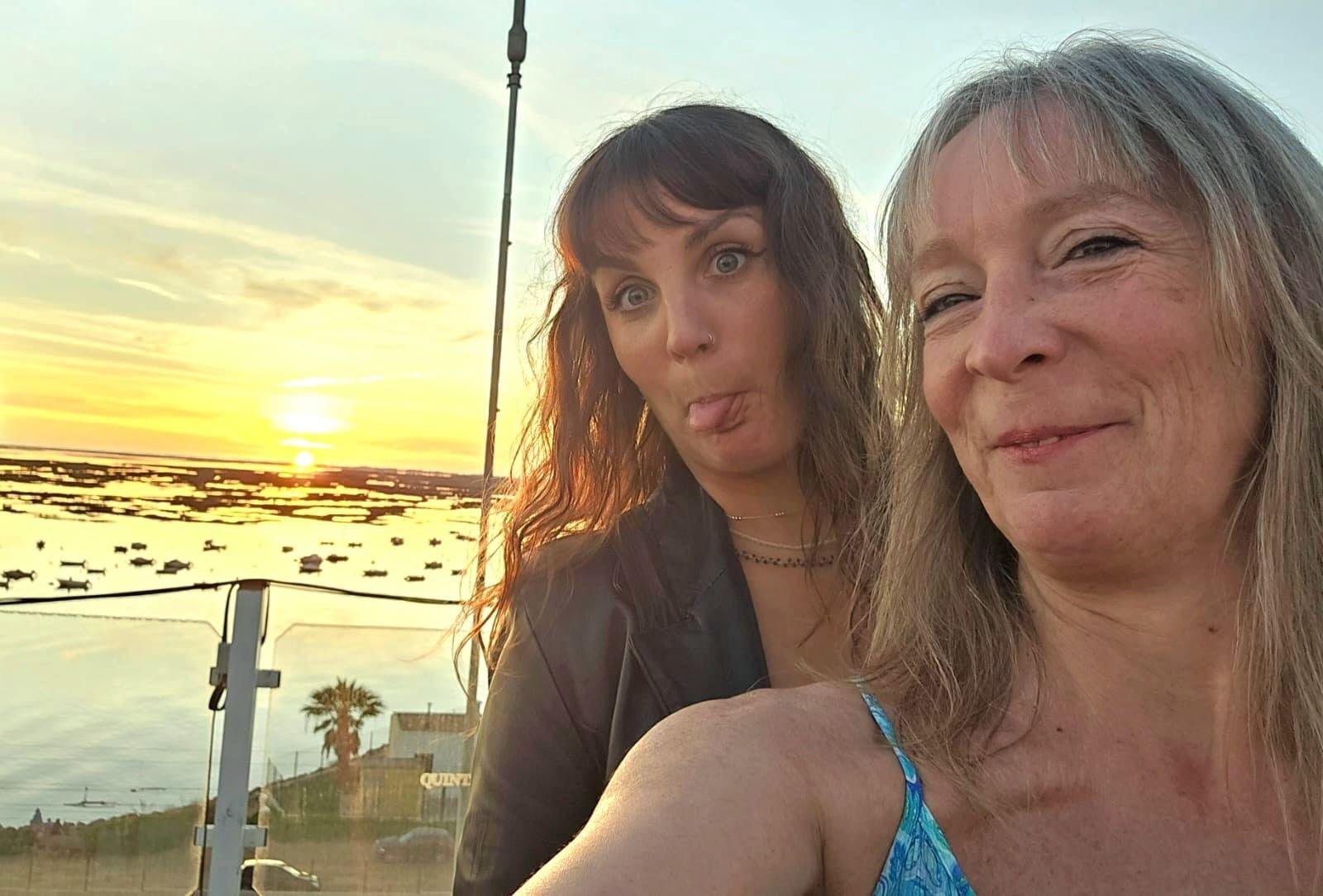 A mother and daughter taking a selfie as the sun sets on a beach. The daughter is sticking her tongue out and the mother smiles at the camera