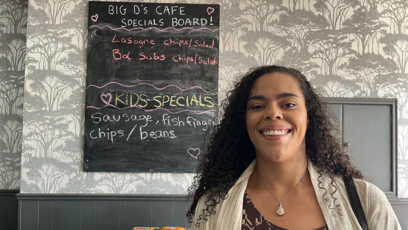 A woman with dark curly hair is smiling standing in front of a chalkboard with the cafe's menu on it. It includes a 'kids special of fish, chips and beans'.