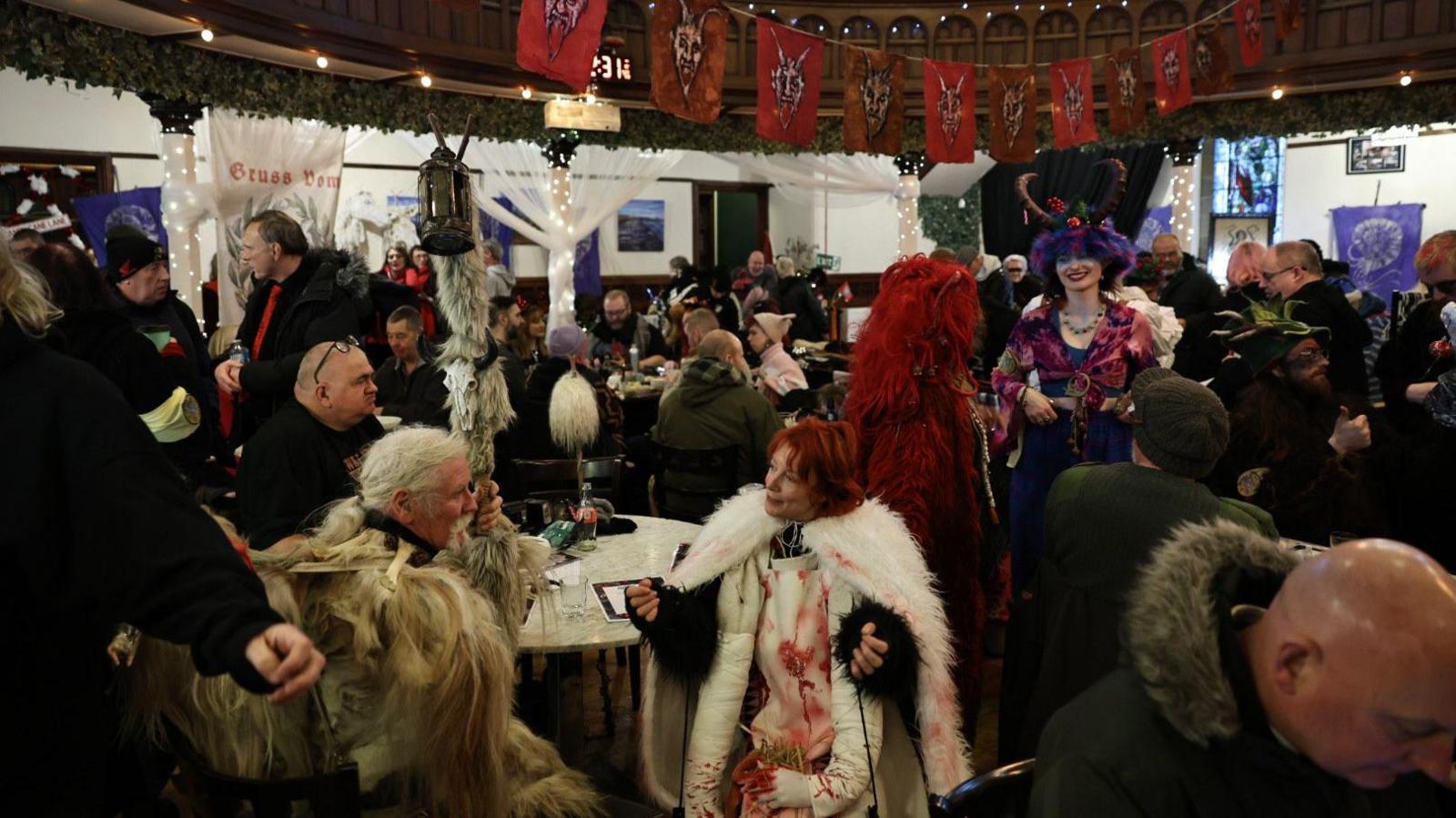 A room filled with people sat at tables celebrating the Krampus festival, some dressed up in fur with horns and blood splatters 