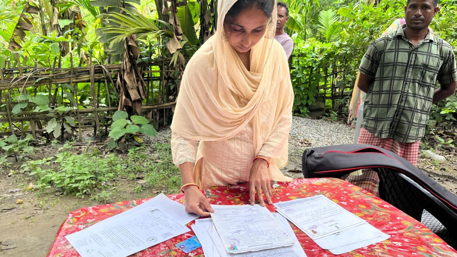 A woman shows papers placed on a red table, she is wearing a pale yellow Indian suit with a scarf in matching colours. A man is standing behind wearing a green shirt.