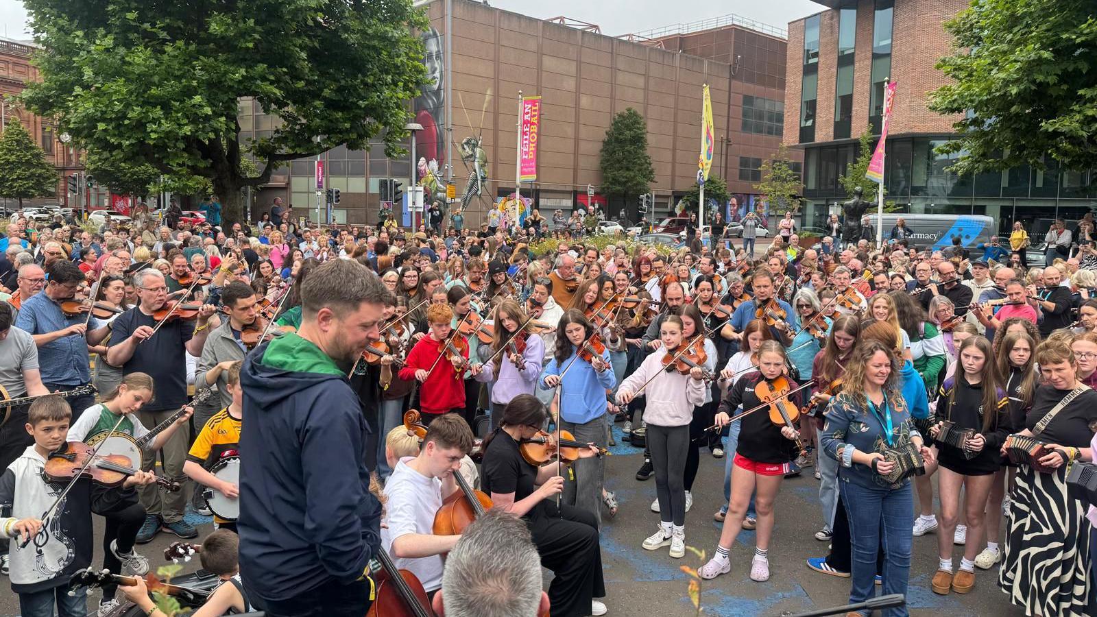 Hundreds of musicians young and old stand in a city centre park, playing a variety of traditional instruments as they are guided by one man in the foreground.