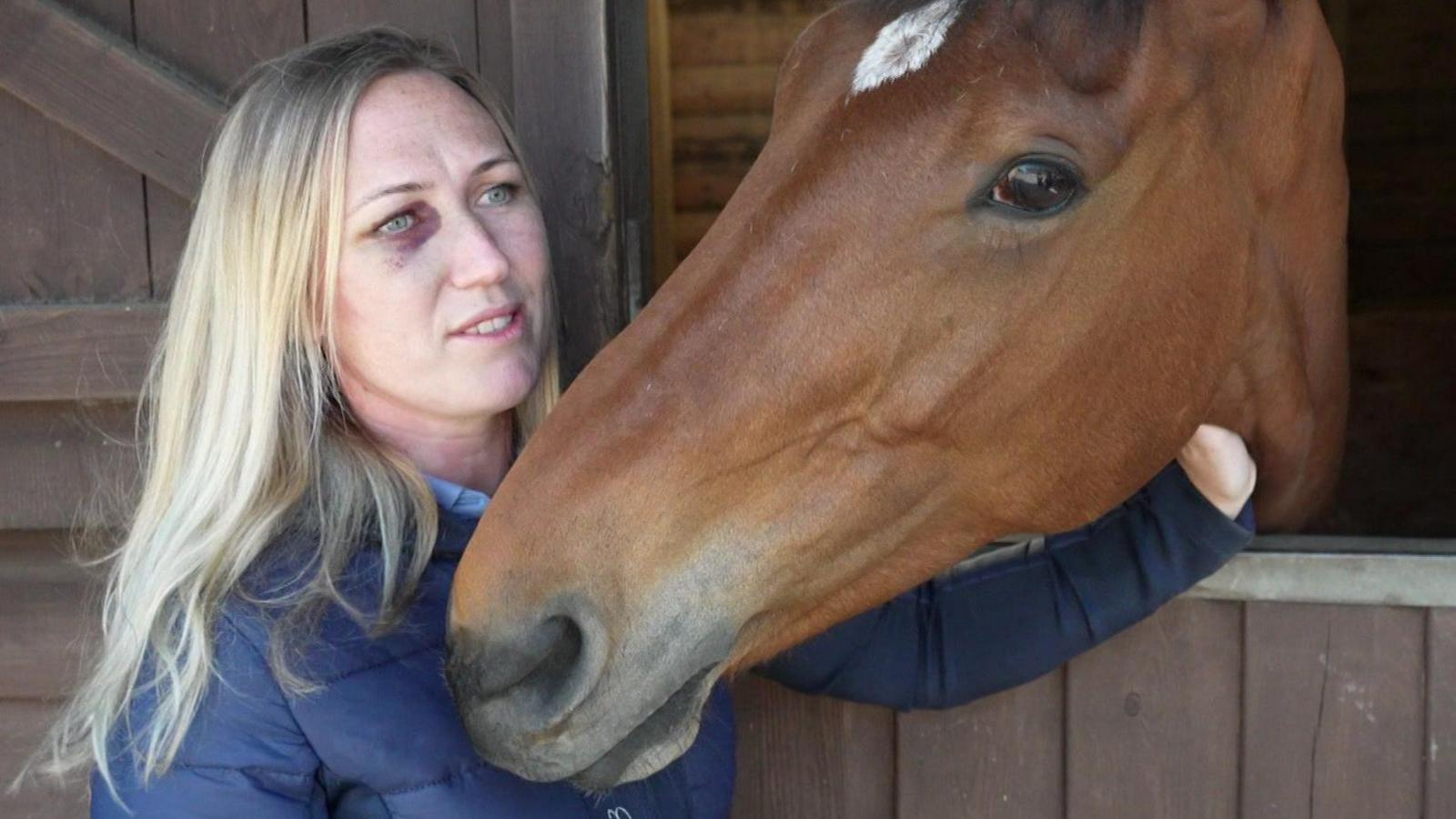 Becci stood next to a stable door with Barney - a brown horse - sticking his head out. Becci, who has long blonde hair and is wearing a blue coat, has a black eye from when she was knocked off Barney. 