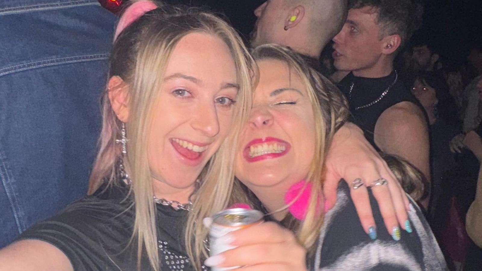 Abi takes a selfie using a digital camera. She smiles widely at the camera. She has long blonde hair and wears a black crop top with a blue jumper tied round her waist. She has her arm around another woman who has long blonde hair and smiles widely at the camera. She wears a black T-shirt and holds a can of drink.