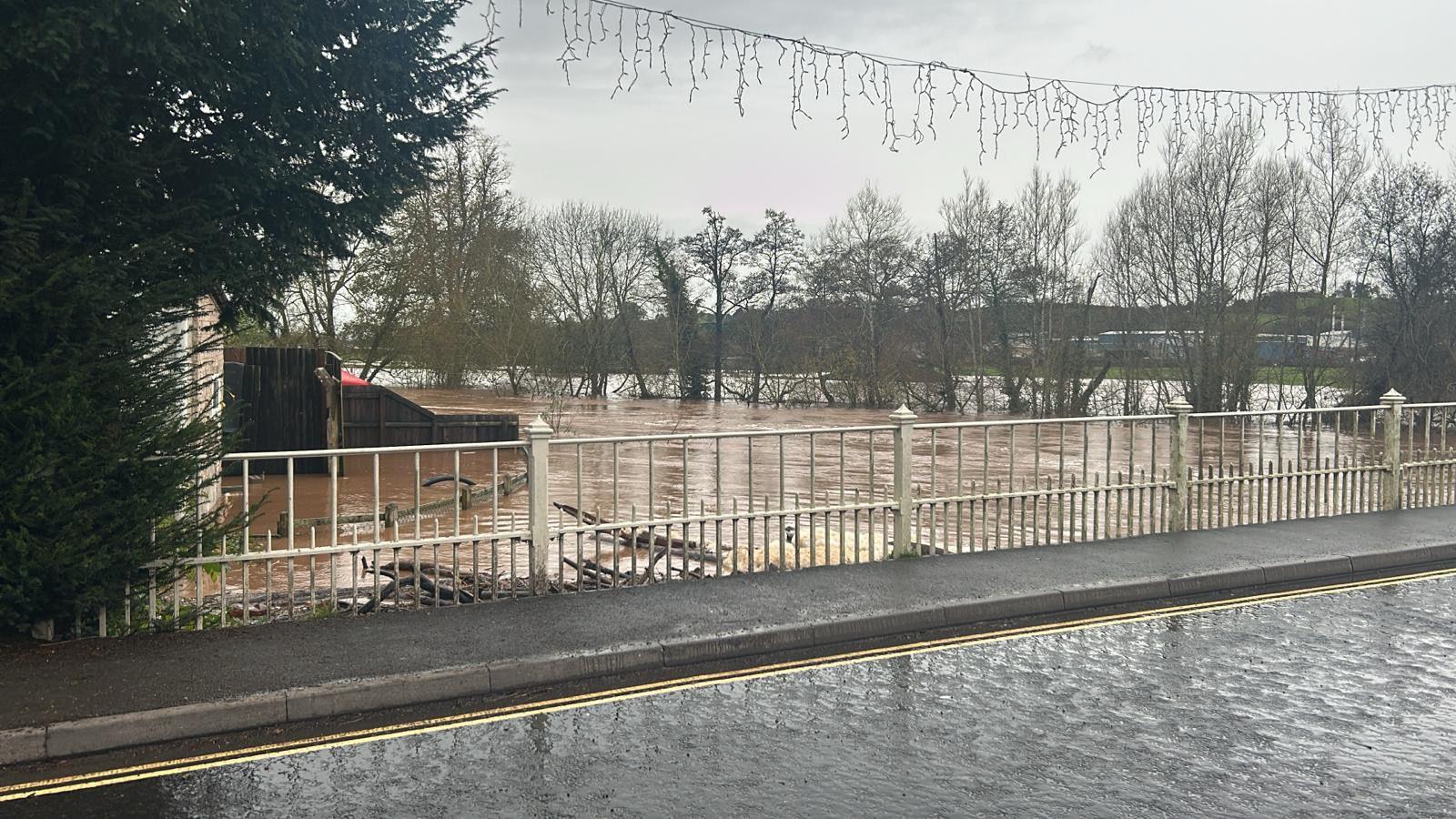 A bridge over a river, which is almost as high as the road. There are white iron railings on the bridge's edge, beyond which brown water can be seen, with submerged trees on the river bank. Further flooding can be seen on surrounding fields.