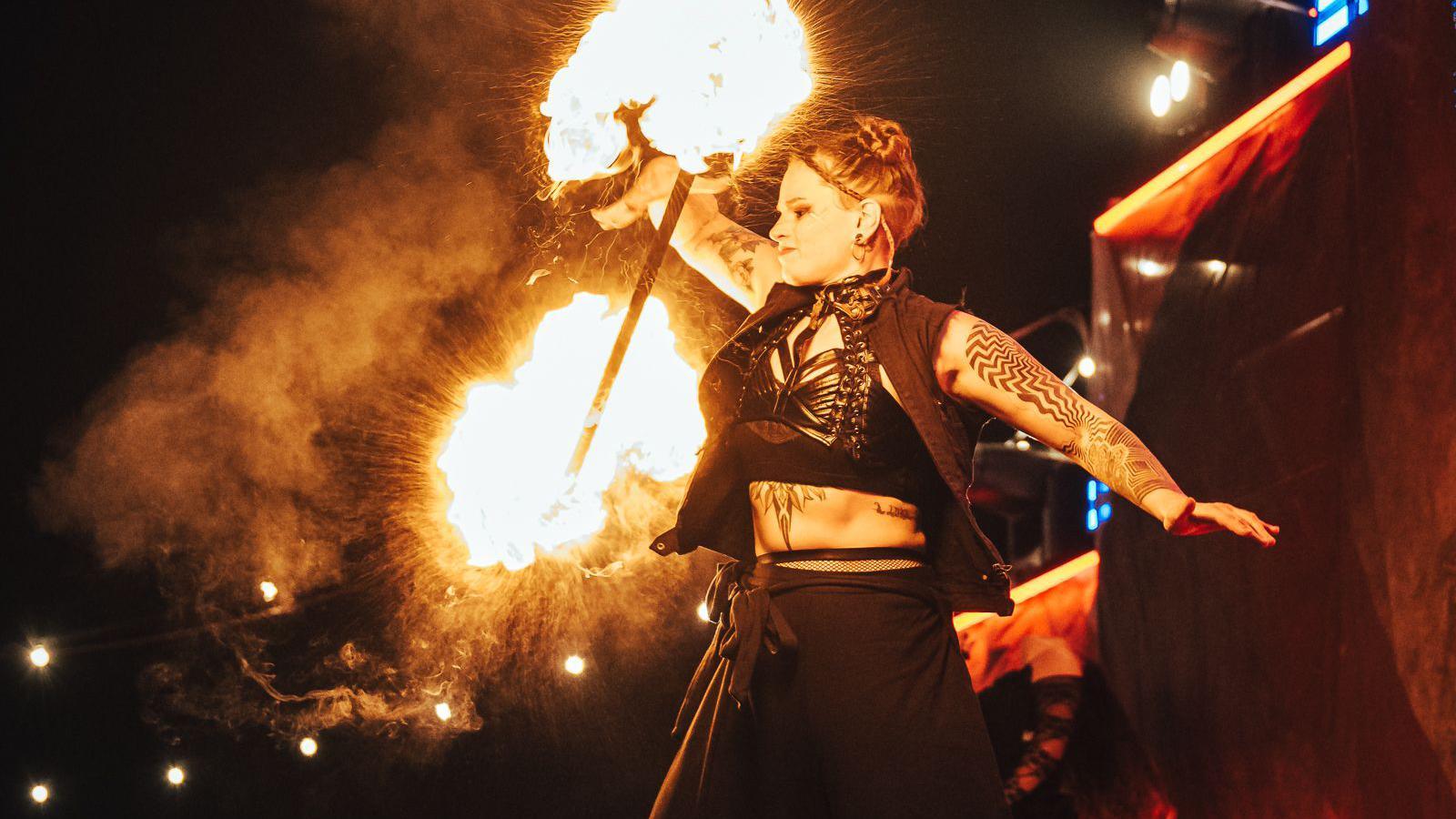 A female fire performer standing on stage as she holds up a baton lit with fire either end in one of her hands. She has tattoos and is wearing a black outfit. The photo has been taken at night.