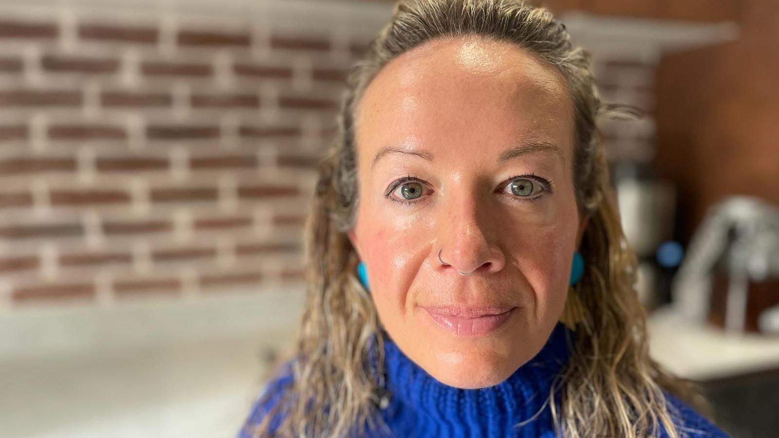 Laura is a white woman with long, fair wavy hair. She is wearing a blue jumper and is stood in the kitchen of her home.