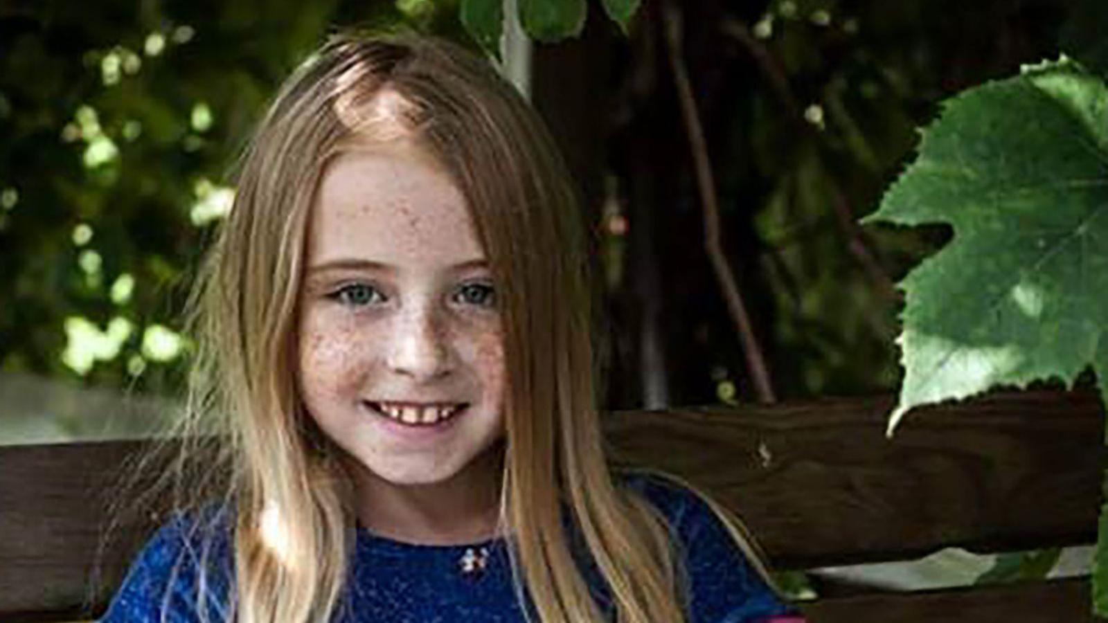 Cimarron Thomas, a young girl with blonde hair smiling at the camera. She has freckles on her face and is wearing a royal blue top. She is sitting on a bench.