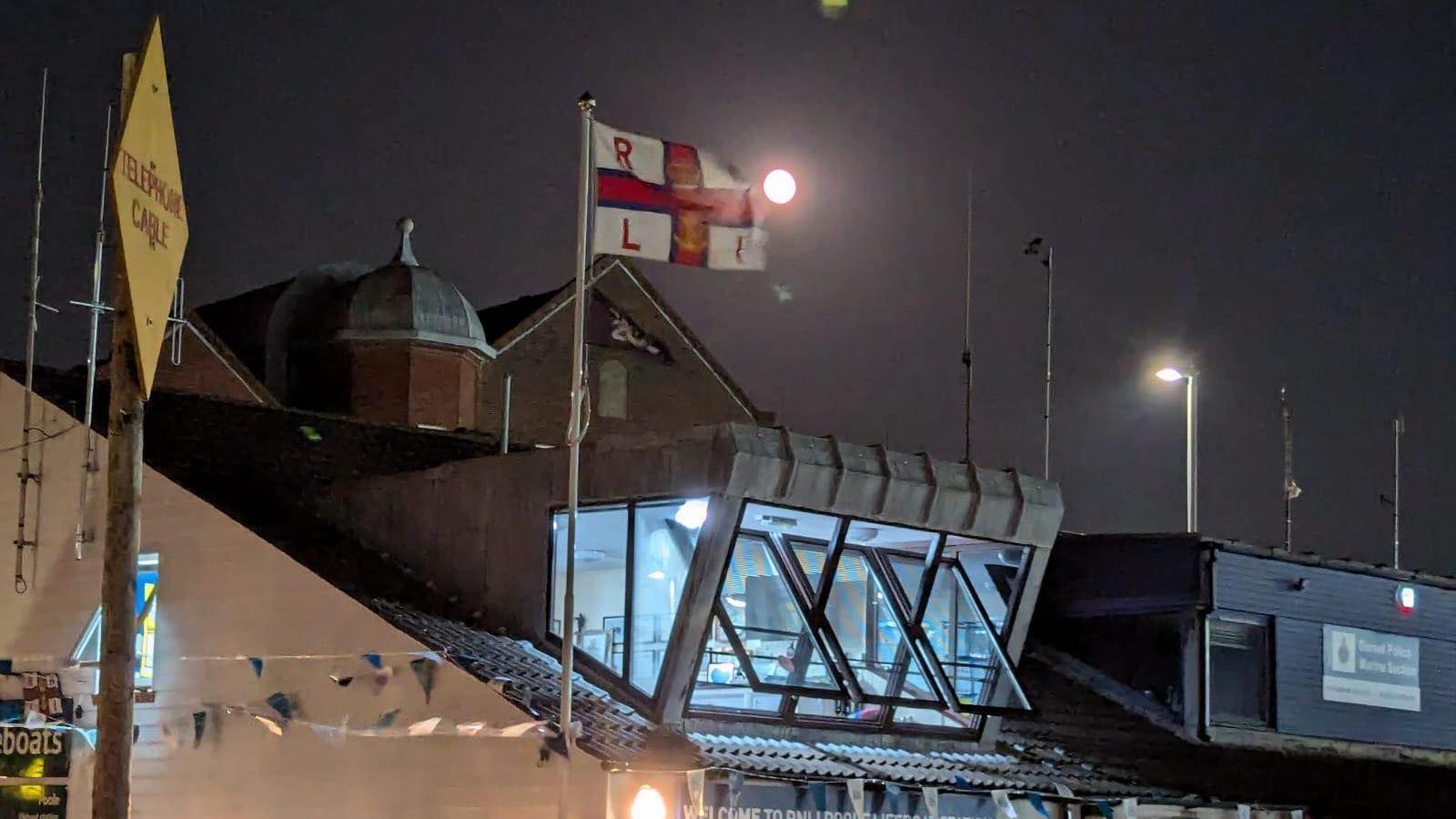 Poole RNLI lifeboat station at night. The red and white RNLI flag is flying on the flagpole and the moon is shining in the sky.