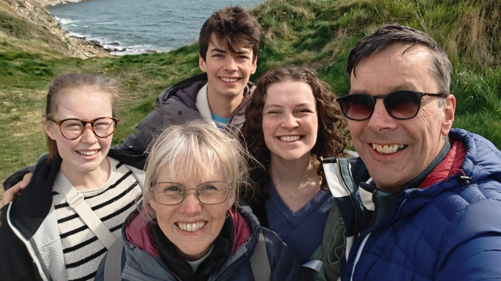 Paul and his family - wife, two daughters and son - all smiling while they are out for a hike with cliffs and the sea in the background