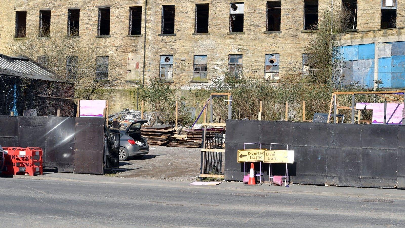 The entrance to a site with two road signs that read "Diverted traffic" to the right. A car with its boot open is parked on the inside, to the left of the entrance and several wooden pallets can be seen stacked at the back.