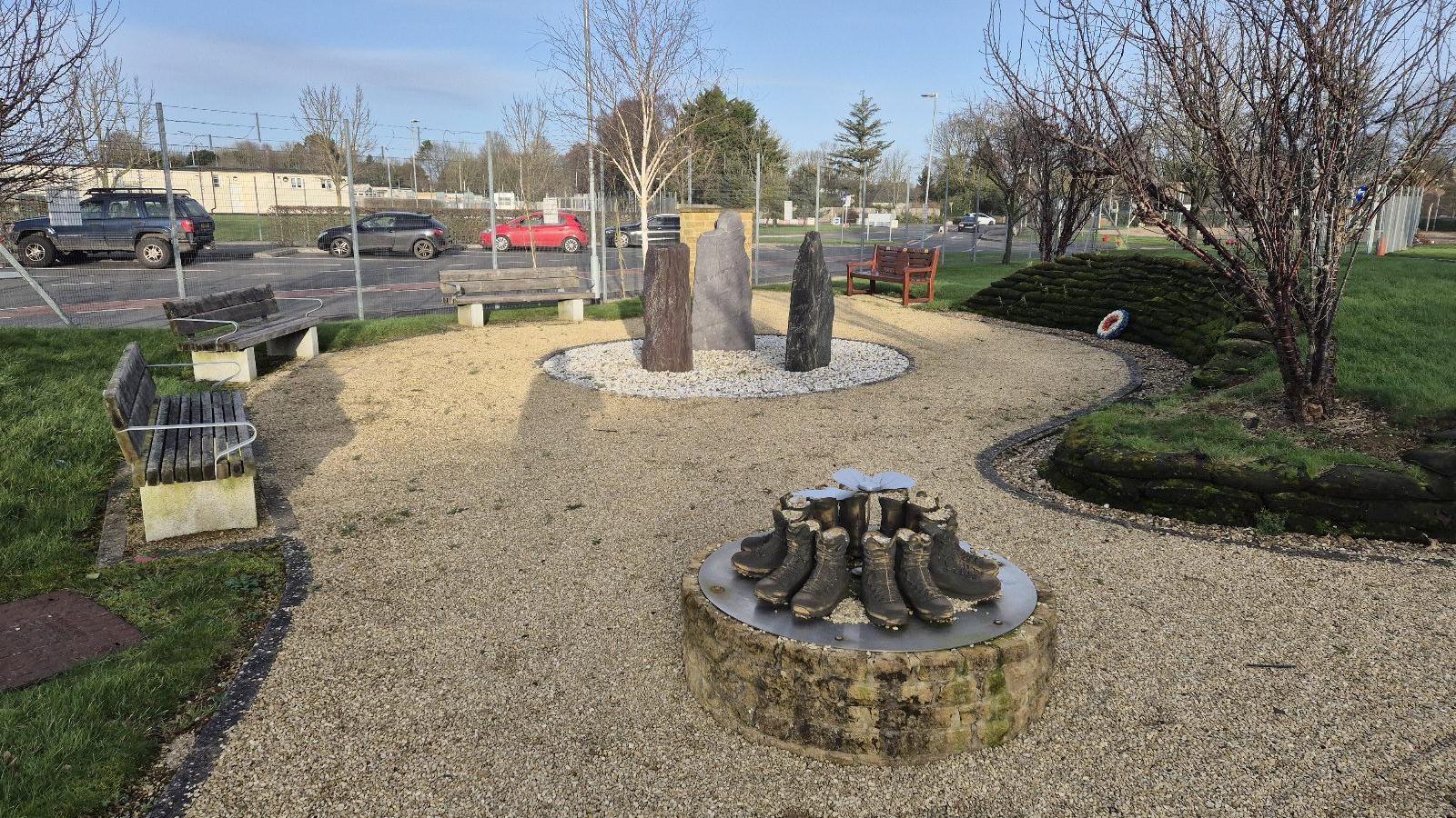 The remembrance garden in Lyneham. There are several benches around a gravelled area. There are three large standing stones in the middle and a separate circular sculpture of boots. A stepped green wall can be seen on one side of the garden and there is a red, white and blue wreath on the ground in front of it.