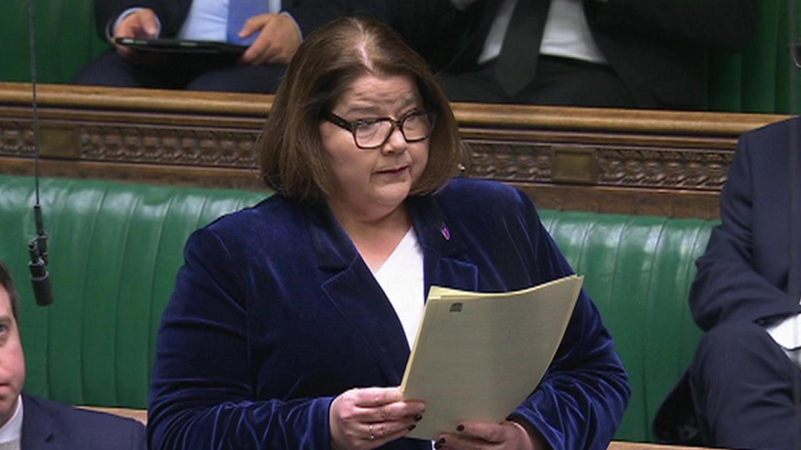 Julie Minns MP is standing in the House of Commons chamber delivering a speech. She is wearing a blue jacket and holding some papers.