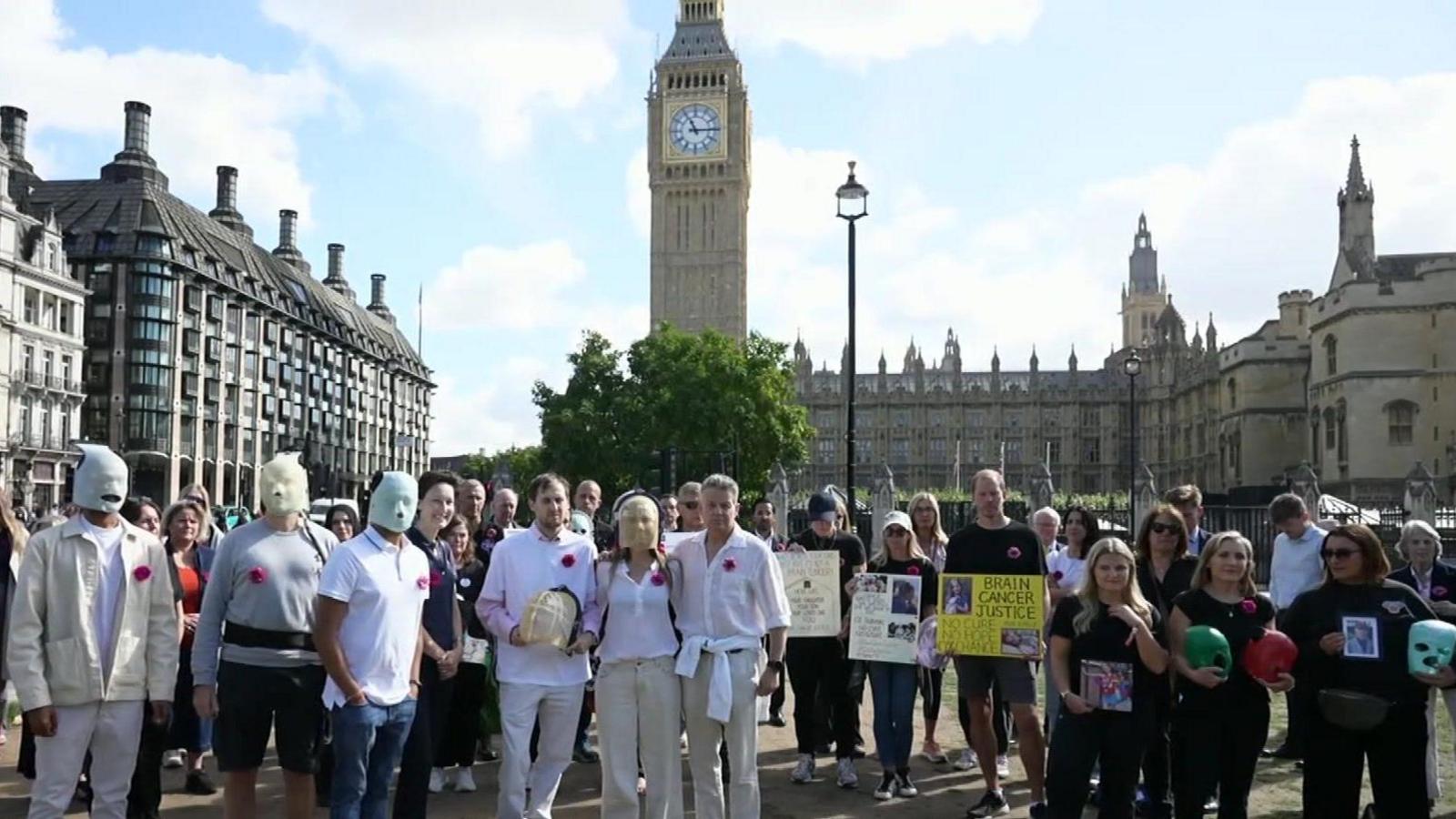 Campaigners for more research into brain cancer gathered by the Houses of Parliament. They are wearing red flowers on their tops. Some are holding placards and some area wearing masks. The Elizabeth Tower can be seen behind them. It is a sunny day.