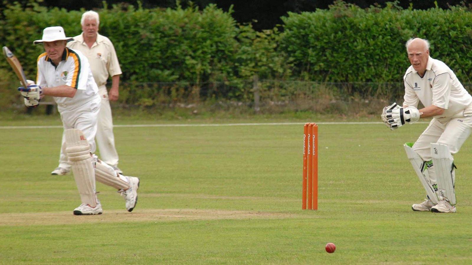 Senior men playing cricket in their whites. You can see orange stumps, a red cricket ball and a batsman with his bat in the air, after hitting the ball on to the on-side. Wicketkeeper Gerald Cooke is leaning forward while looking at the ball. A fielder is in the background.