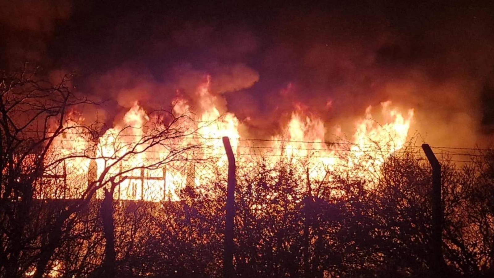 A large fire can be seen coming from a building at night time. A fence can be seen in front of the blaze and smoke billows into the night sky.