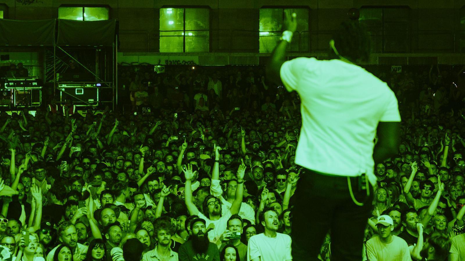 The lead singer of Bloc Party gestures to a large crowd at the Siren Bristol festival in Bristol. He is standing on the stage looking out over the Lloyds Ampitheatre and the picture is taken from behind him. The crowd are bathed in green light