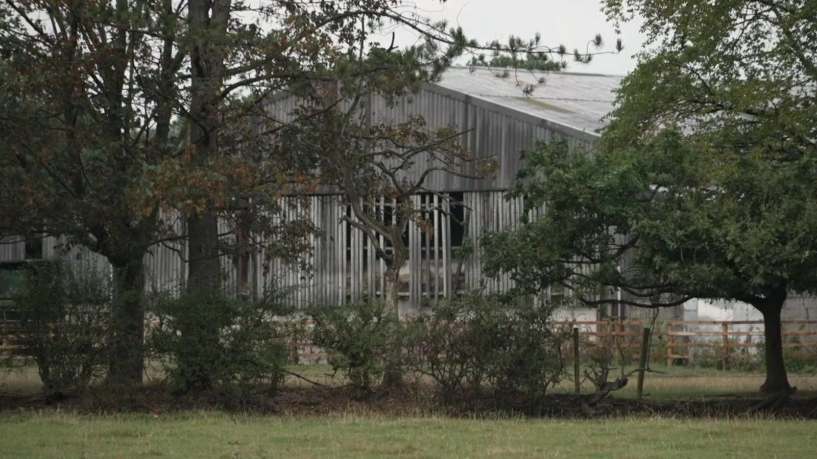 Exterior shot of a farm building. In the foreground you can see a grassy area with large trees. The grey farm building is in the background. 
