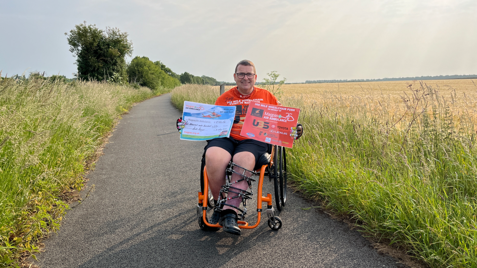 A man in a wheelchair on the path where he started the challenge. The concrete path has overgrown grass on both sides. He is wearing glasses, has short brown hair and is smiling at the camera. His wheelchair has black wheels and an orange frame. His left leg is encased in an external cage.