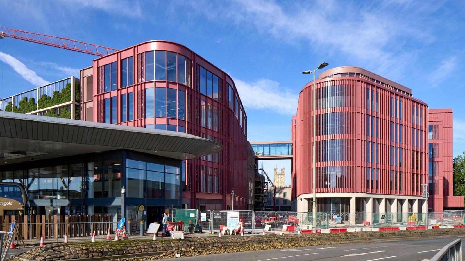 The Forum in Gloucester as seen from the railway station side of Bruton Way, opposite Gloucester Transport Hub. The Forum is a red brick modern building which has a glass walkway linking to another part of the building. Gloucester Cathedral can be seen in the distance, underneath the walkway.